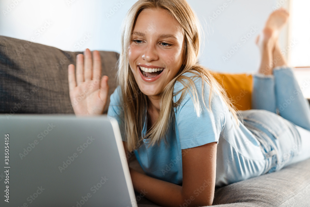 Charming happy girl waving hand and using laptop while lying on couch Charming happy girl waving hand and using laptop while lying on couch