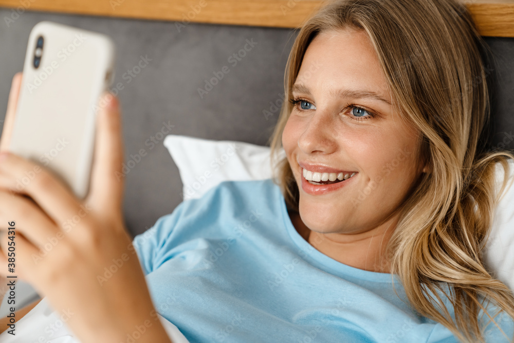 Charming smiling blonde girl using cellphone while lying on bed Charming smiling blonde girl using cellphone while lying on bed