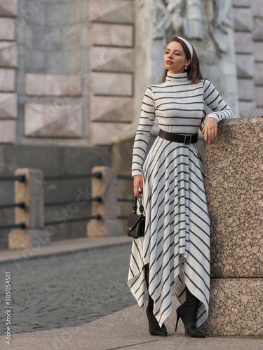 Elegant woman in long warm dress with grey and white stripes walking at city street. Female model with long wavy brunette jair in sunglasses with black leather handbag photo