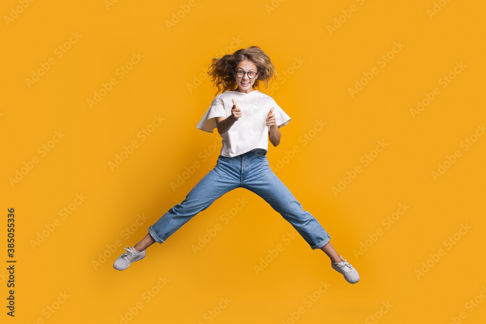Jumping blonde woman is gesturing the shot and gun with both hands while jumping on a yellow wall at studio