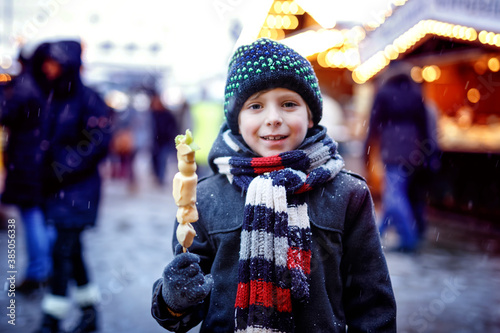 Little cute kid boy eating white chocolate covered fruits on skewer on traditional German Christmas market. Happy child on traditional family market in Germany during snowy day.