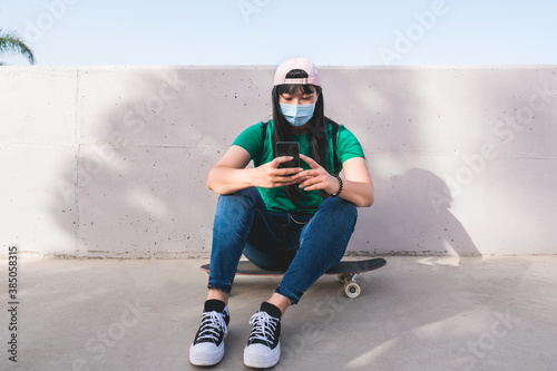 Beautiful asian girl sitting on skateboard and looking at smartphone while wearing surgical face mask - Coronavirus lifestyle
