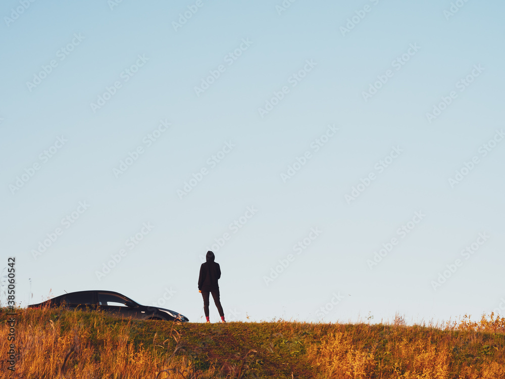 Obraz premium A girl in black clothes stands next to a black sedan car on a hill, against a dim sky, copy space. Concept of auto tourism and travel. Silhouette of a girl and a car at sunset in autumn