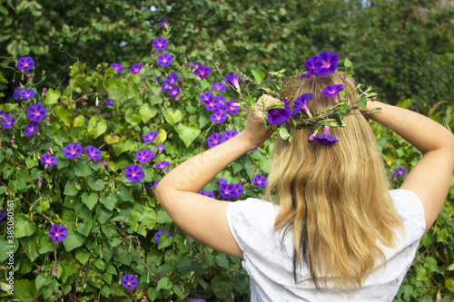 a woman against the background of nature holds a wreath of blue flowers on her head with two hands, against the background of bushes with blue flowers