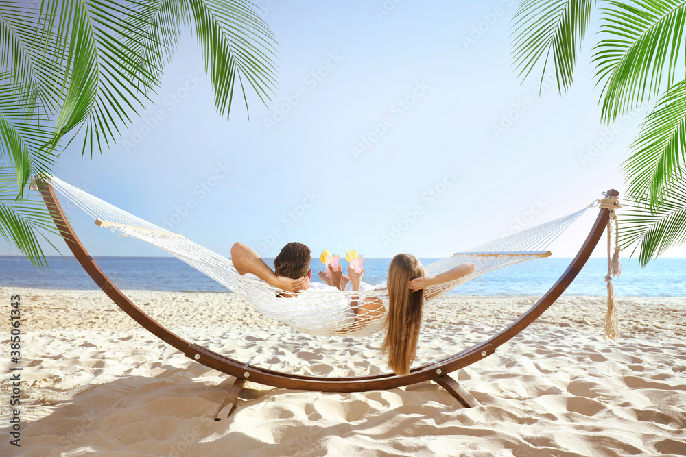 Couple with refreshing cocktails relaxing in hammock under green palm leaves on beach - obrazy, fototapety, plakaty 