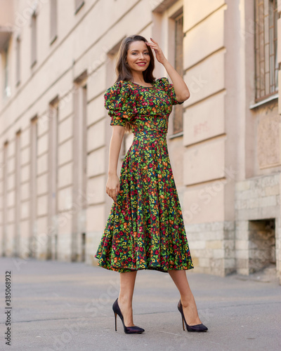 Elegant caucasian brunette woman with long wavy hair in midi green dress with floral print walking at city street on autumn day
