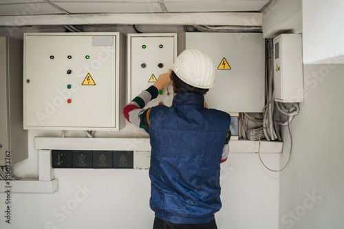 An electrician in an engineering helmet opens the switch box. The technician works in the electrical control room. Service of power distribution switchboards. Back view photo