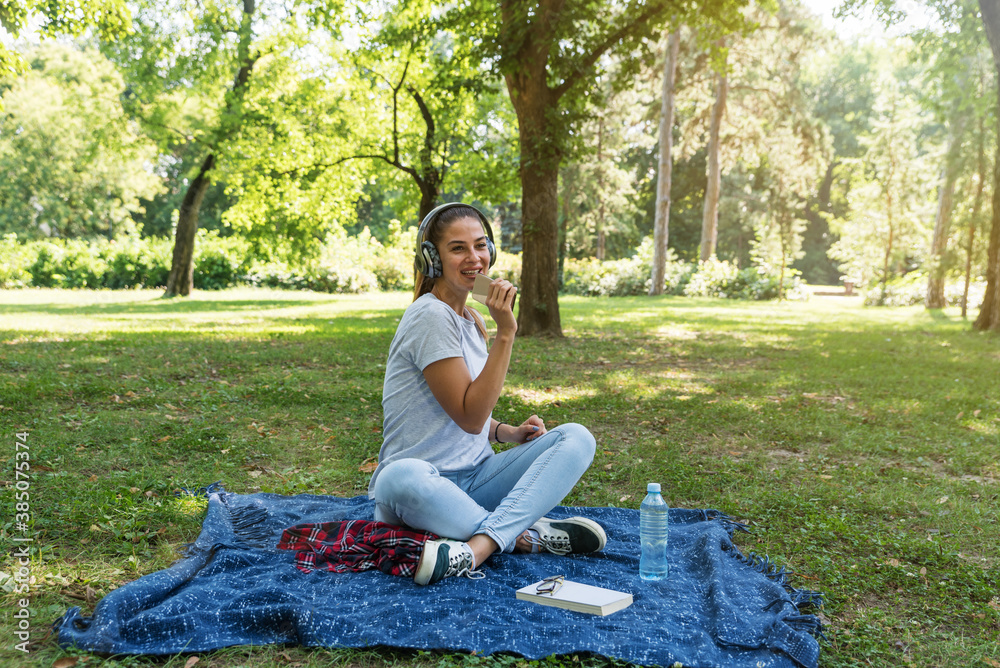 Young woman sitting in the park surfing the song play list in her phone and listening the music on the wireless headphones Young woman sitting in the park surfing the song play list in her phone and listening the music on the wireless headphones