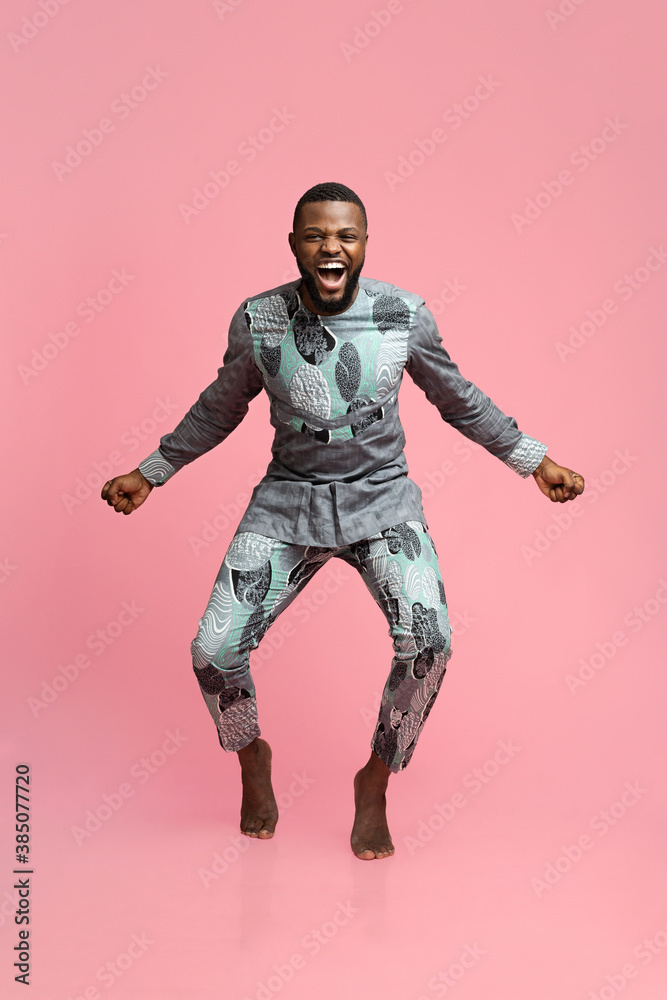 Emotional black guy in african costume dancing over pink background