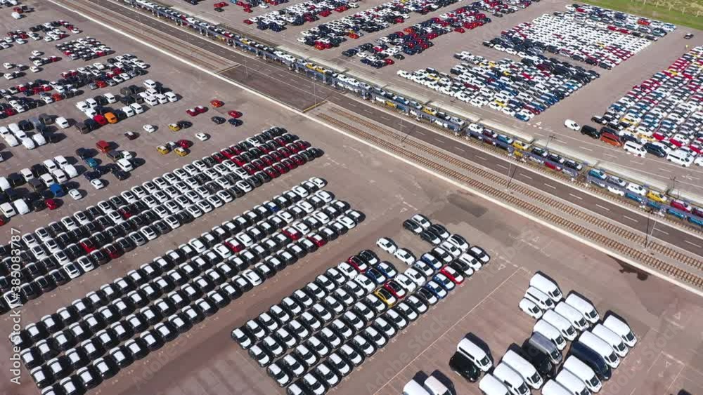 Aerial - Automotive industry. Flying over port full of cars and vans. Train leaving port with stocked cars Aerial - Automotive industry. Flying over port full of cars and vans. Train leaving port with stocked cars