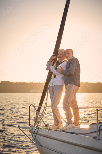 Full leghth of beautiful happy senior couple in love standing on the side of sail boat floating in sea at sunset, they are hugging and enjoying amazing evening view photo