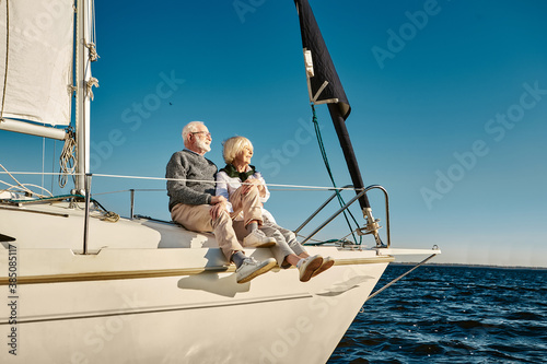 Enjoying vacation. Happy senior family couple sitting on the side of a sail boat or yacht deck floating in a calm blue sea, hugging and enjoying amazing view