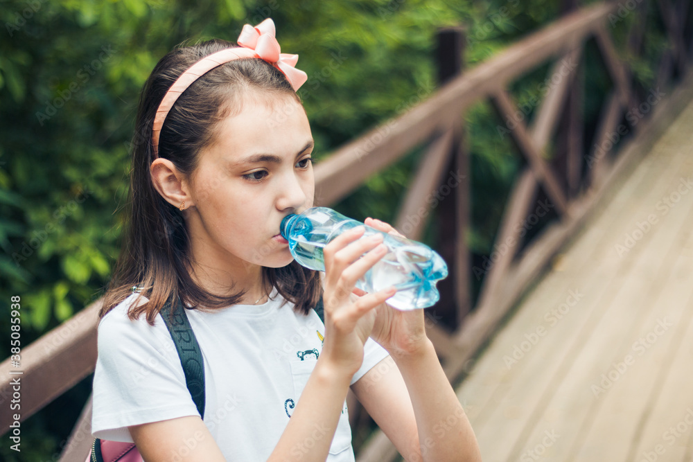Portrait of a charming child holding a bottle of water, in t-shirt with a backpack on his back, standing in the open air Portrait of a charming child holding a bottle of water, in t-shirt with a backpack on his back, standing in the open air