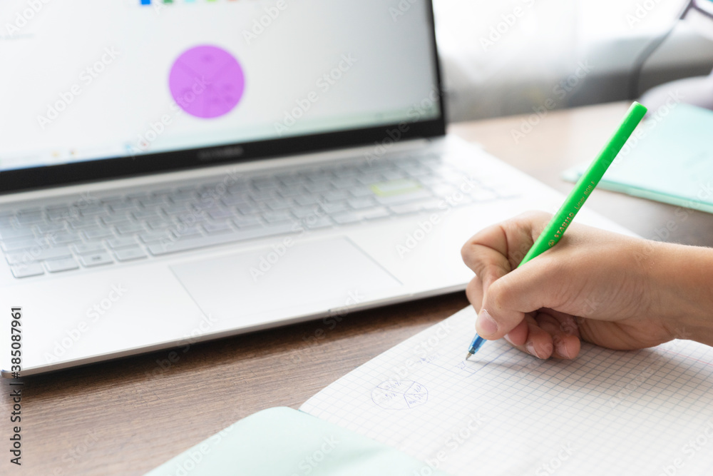 child's hands write homework in school notebook, laptop with screen with charts stand near on table. Remote home education concept. New reality child's hands write homework in school notebook, laptop with screen with charts stand near on table. Remote home education concept. New reality