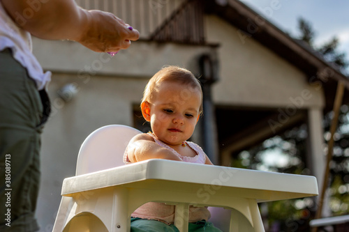 Shoto of a baby girl eating porridge. Mom feeding daughter with a spoon. Mother feeding her cute baby girl outdoor in the nature while sitting in the high baby chair. Baby has breakfast in highchair. photo