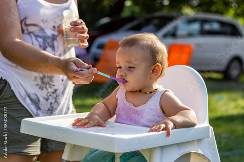 Shoto of a baby girl eating porridge. Mom feeding daughter with a spoon. Mother feeding her cute baby girl outdoor in the nature while sitting in the high baby chair. Baby has breakfast in highchair. photo