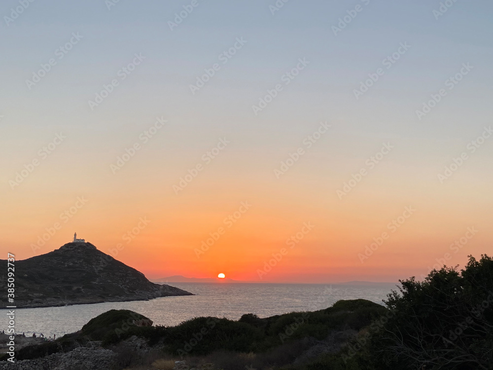 Beautiful romantic sunset on the Aegean Sea with a lighthouse on the top of a hill. Reflections of the dawn over the sea. Ruins of some ancient stones. 