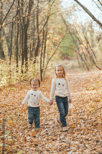 Portrait of two cheerful small sisters in white sweaters and jeans walking in the park in warm autumn day