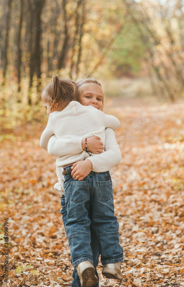 Fototapeta premium Two cheerful small sisters in white christmas sweaters and jeans hugging in the park in warm autumn day
