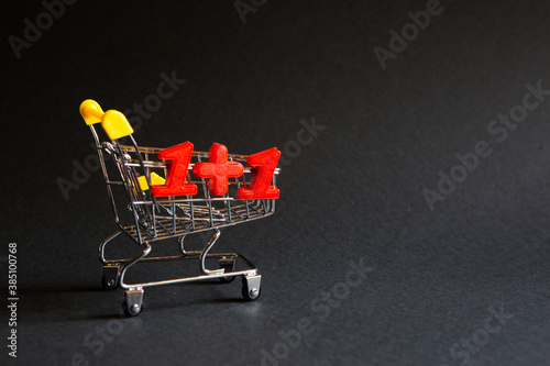 Shopping cart and the numbers "1+1" on a black background. Black Friday, discounts, sale, shopping, two for the price of one, one plus one. Space for text