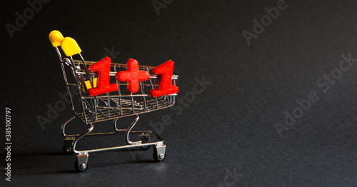 Shopping cart and the numbers "1+1" on a black background. Black Friday, discounts, sale, shopping, two for the price of one, one plus one. Space for text