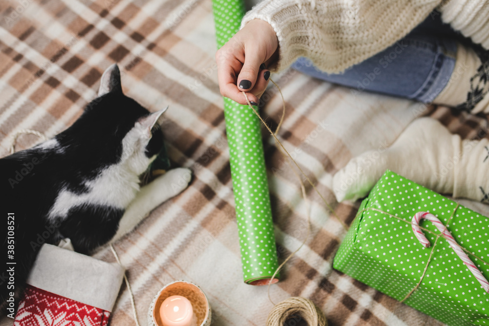 Φωτογραφία Young woman sits on plaid in cozy knitted woolen white sweater, socks and wraps Christmas gift in polka dot wrapping paper