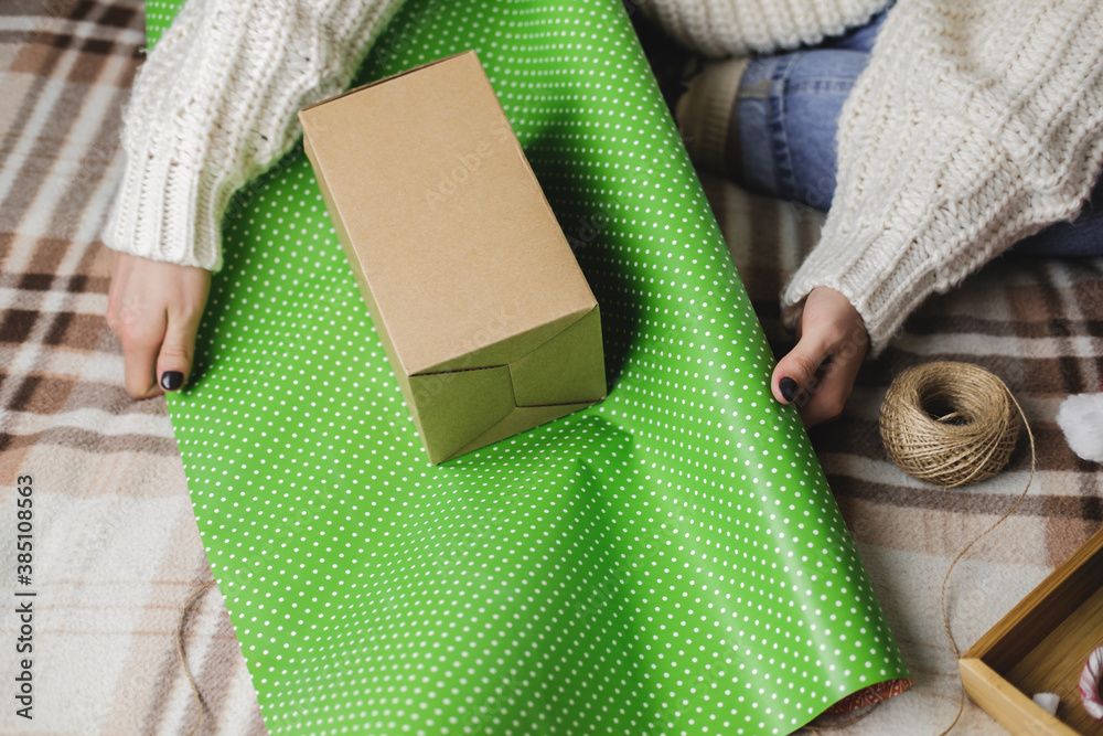 Φωτογραφία Young woman sits on plaid in cozy knitted woolen white sweater, socks and wraps Christmas gift in polka dot wrapping paper