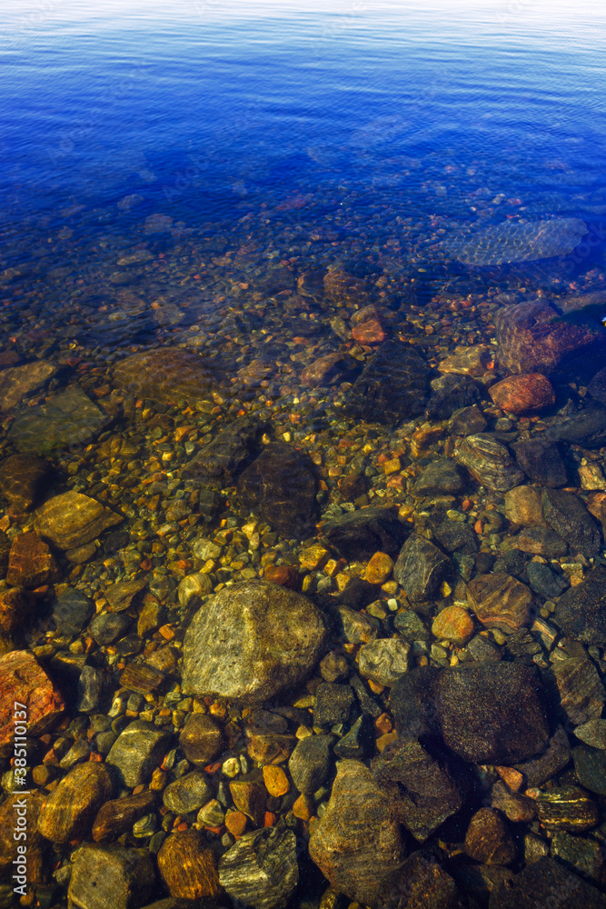 Stones at the bottom of a clear lake with clear water with a blue sky reflection.