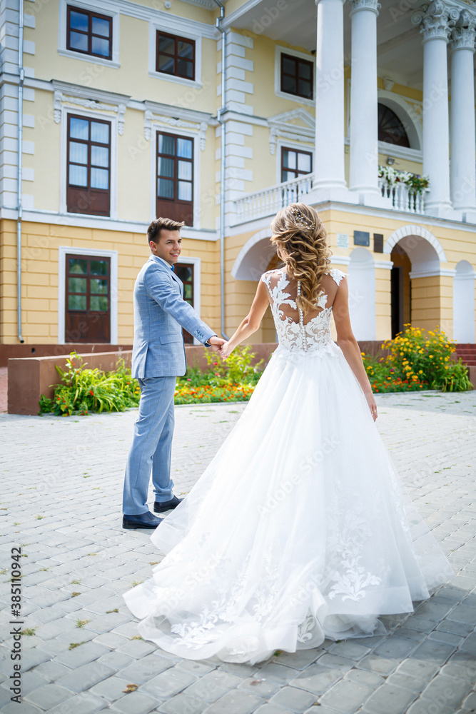 Naklejka premium Young stylish guy in a suit the groom and the bride beautiful girl in a white dress with a train walk in the park on their wedding day