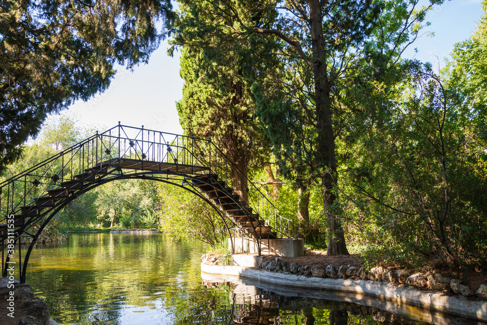 Horizontal view of a metal bridge over a pond with trees, in El Capricho park, in Madrid, Spain Horizontal view of a metal bridge over a pond with trees, in El Capricho park, in Madrid, Spain