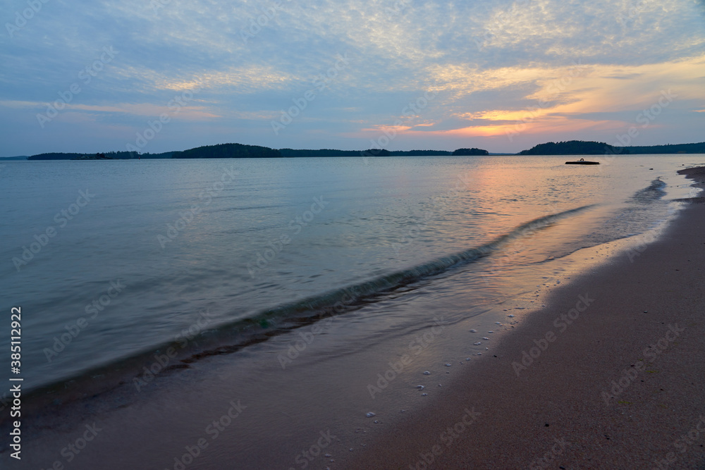 Sunset on a sandy beach with reflection of sky on water surface and waves hitting sand.
