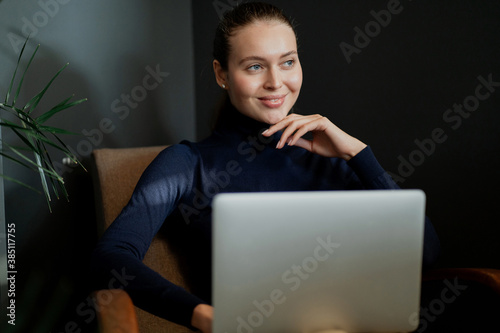 successful young woman of European appearance works on a laptop computer, sits in a comfortable chair, modern clothes for the office. Thinks about a successful career in business