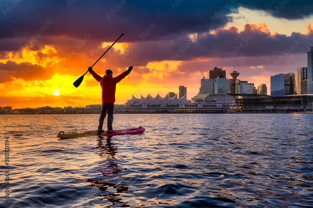 Fototapeta premium Adventurous man is paddle boarding near Downtown City during a vibrant winter sunrise. Dramatic Sky Artistic Render. Taken in Coal Harbour, Vancouver, British Columbia, Canada. Colorful Sky Overlay