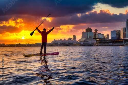 Adventurous man is paddle boarding near Downtown City during a vibrant winter sunrise. Dramatic Sky Artistic Render. Taken in Coal Harbour, Vancouver, British Columbia, Canada. Colorful Sky Overlay