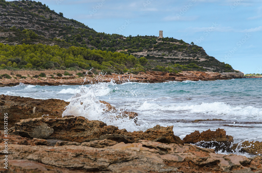 Sea waves break against the rocky coastline with Badum sentinel tower in the background, Castellon, Spain
