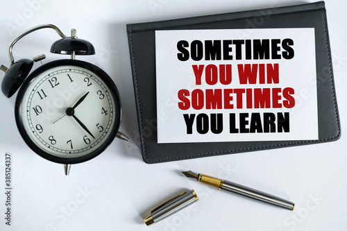 On the table there is a clock, a pen, a notebook and a card on which the text is written - SOMETIMES YOU WIN SOMETIMES YOU LEARN