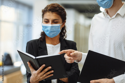 Two collegues discuss work and business wearing protective sterile medical masks standing in the corridor of the office. Coronavirus spread prevention during global pandemic concept.