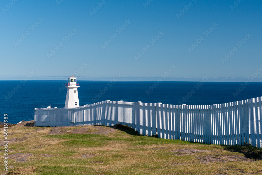 A white circular lighthouse with a green beacon light. The building has multiple windows. There's a long white fence in front of the tower with deep blue ocean in the background and a bright blue sky.