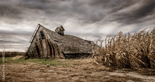 An old, wooden corn crib collapsed under its own weight. The barn is surrounded by a field of corn, ready for harvest as a storm clouds gather. Concepts of family farms, heartland, midwest