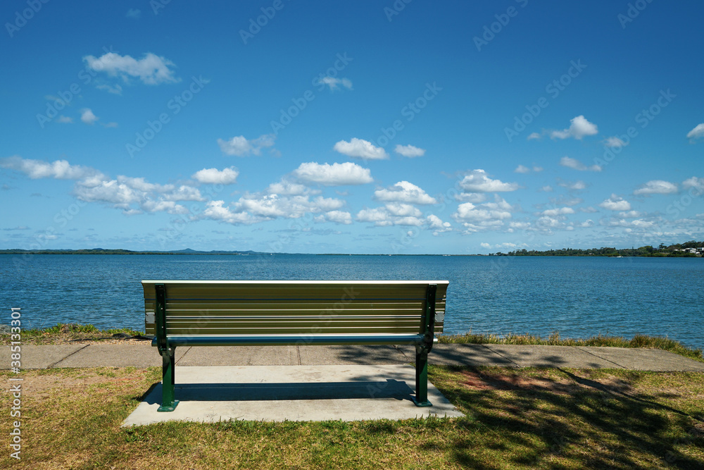 A place to sit and relax. Bench seat beside the water, with a view across the bay to islands on the horizon, and blue sky with fluffy clouds. Victoria Pt, Moreton Bay, Redlands, Queensland, Australia.