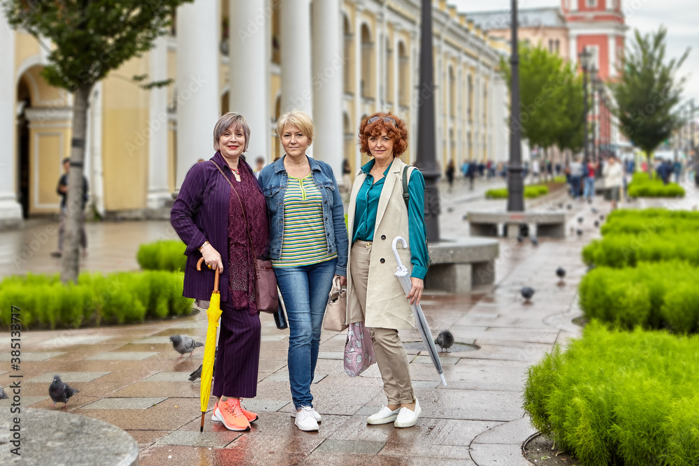 Three mature women stand on a city street in rainy weather and look at the camera.