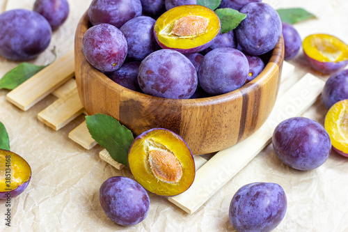 Fresh raw blue and violet plums in wooden bowl with green leaves on light paper background.