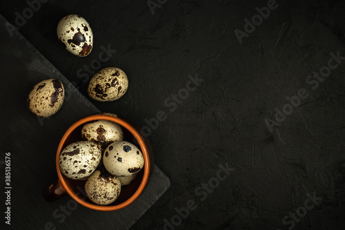raw quail eggs in a clay bowl and on a black stone plate on a textured dark concrete surface. moody artistic mockup with copy space. top view