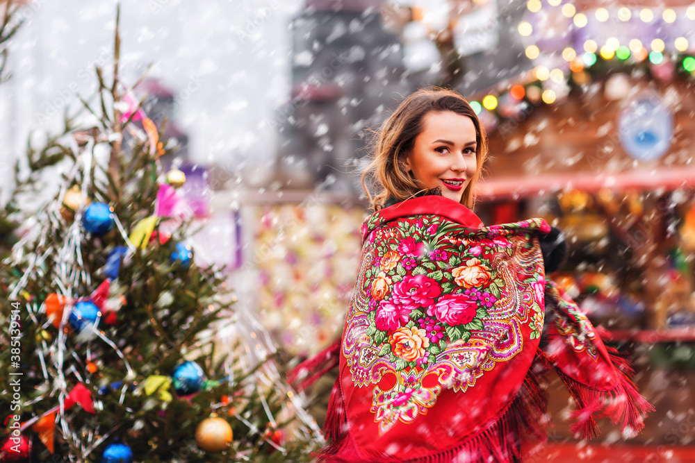 Outdoors lifestyle portrait of pretty blonde young woman. Smiling and walking on the christmas market. Wearing stylish shawl with a floral pattern in the Russian style. It's snowing. Festive mood Outdoors lifestyle portrait of pretty blonde young woman. Smiling and walking on the christmas market. Wearing stylish shawl with a floral pattern in the Russian style. It's snowing. Festive mood
