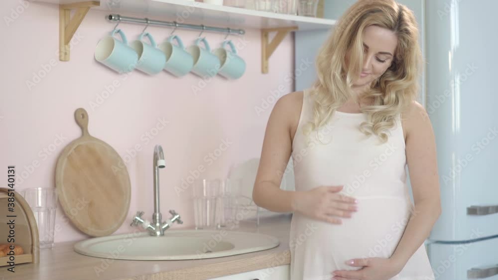 Camera approaches to happy pregnant woman standing in kitchen and stroking belly. Portrait of relaxed confident Caucasian lady enjoying pregnancy at home. Camera approaches to happy pregnant woman standing in kitchen and stroking belly. Portrait of relaxed confident Caucasian lady enjoying pregnancy at home.