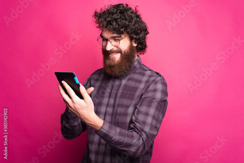 A bearded man is holding a tablet and smiling at it near pink wall