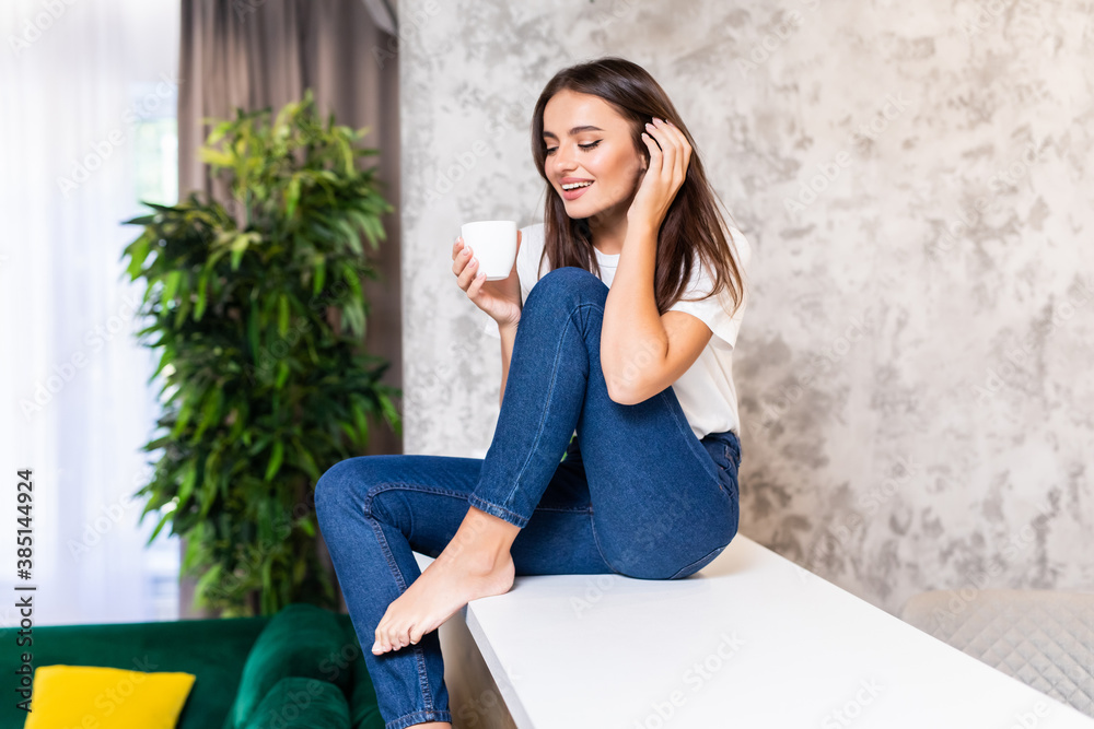 Enjoying time at home. Beautiful young smiling woman working on laptop and drinking coffee while sitting on the countertop in the kitchen at home. Enjoying time at home. Beautiful young smiling woman working on laptop and drinking coffee while sitting on the countertop in the kitchen at home.