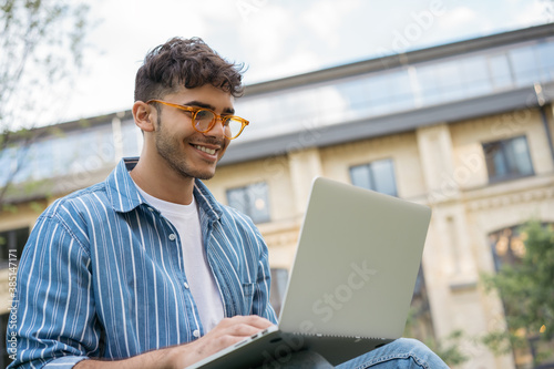 Portrait of happy Indian programmer using laptop computer, internet, working freelance project online, sitting in park. Asian student studying, learning language, using modern technologies outdoors
