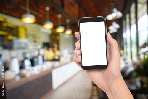 Hand holding smartphone with white blank screen on blurred coffee shop background.