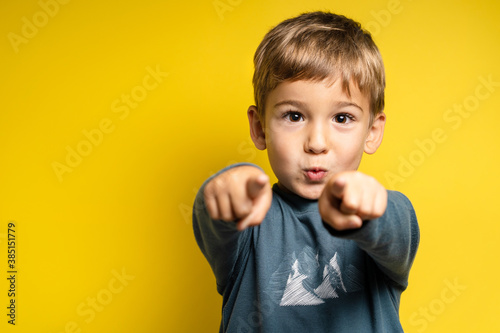 Portrait of happy small caucasian boy in front of yellow background pointing finger - Childhood growing up and achievement concept - front view waist up copy space